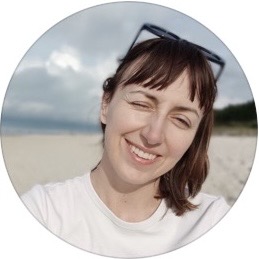 Woman with short brown hair wearing a white shirt and sunglasses on her head, smiling and winking at the beach.