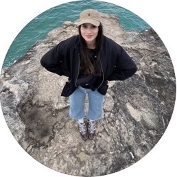 Person wearing a beige cap, black jacket, and blue jeans standing on rocky ground near water.
