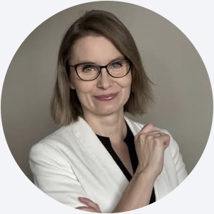 Woman with glasses and shoulder-length hair wearing a white blazer and black top, posing with a slight smile against a plain background.
