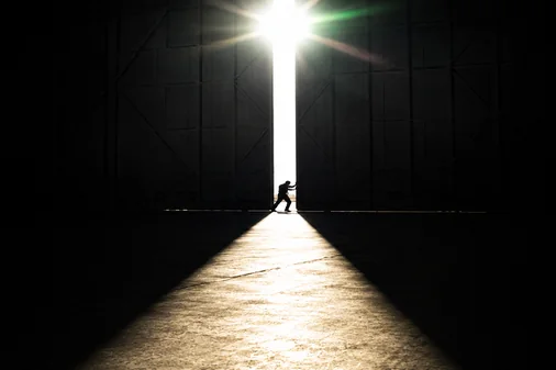 Person opening large industrial doors with bright light shining through in a dark warehouse.
