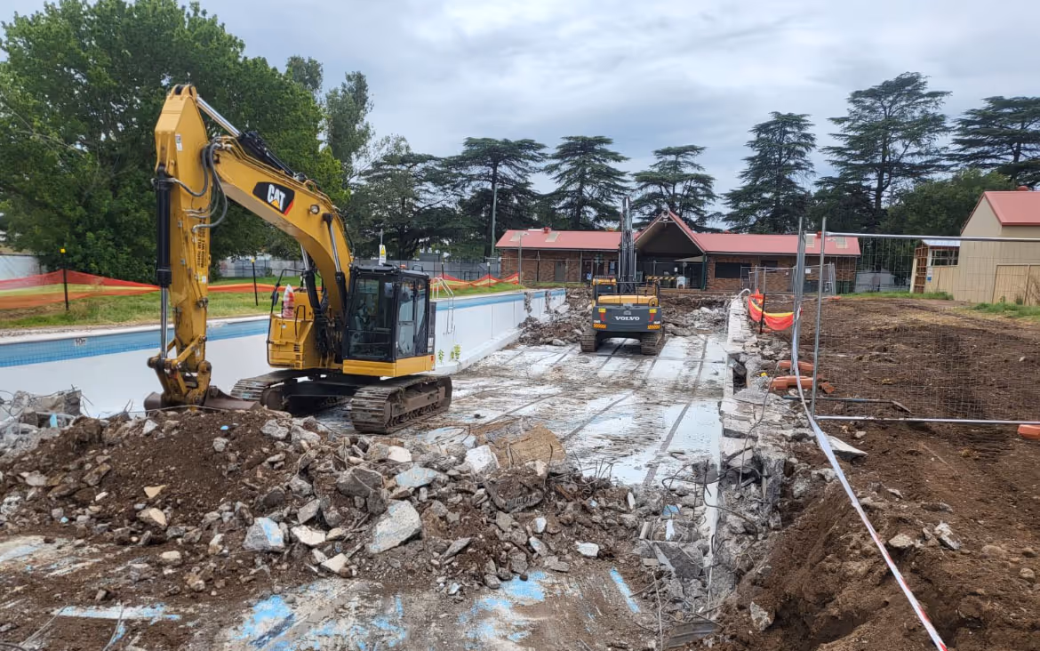 Excavators working inside an empty, partially demolished swimming pool surrounded by construction debris and dirt.
