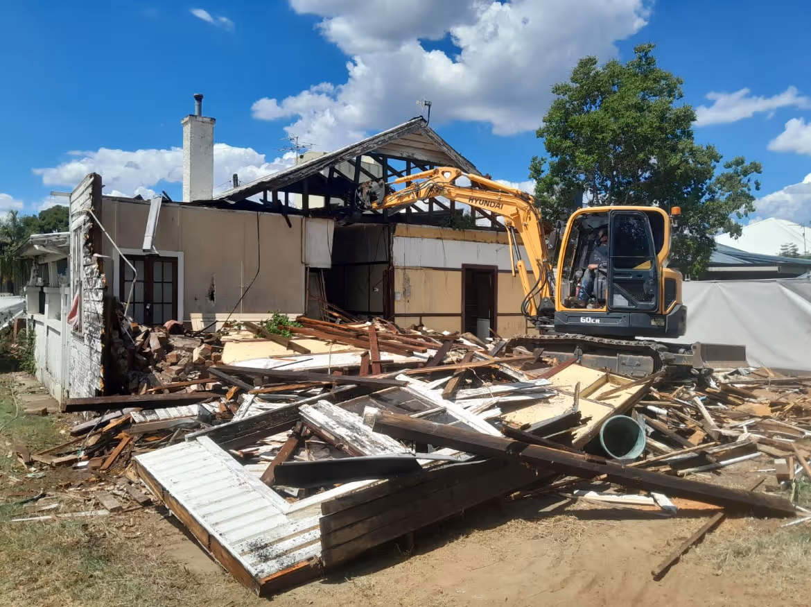 Yellow Hyundai excavator demolishing a partially torn-down house with debris scattered around.