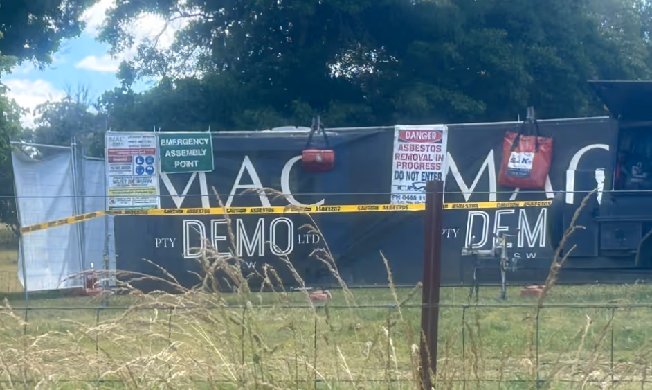 Outdoor asbestos removal site with warning signs, yellow caution tape, and a fenced-off area near a trailer.