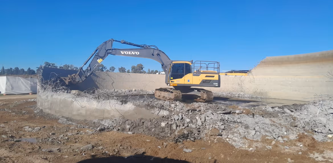Yellow and black Volvo excavator demolishing a large concrete structure under a clear blue sky.