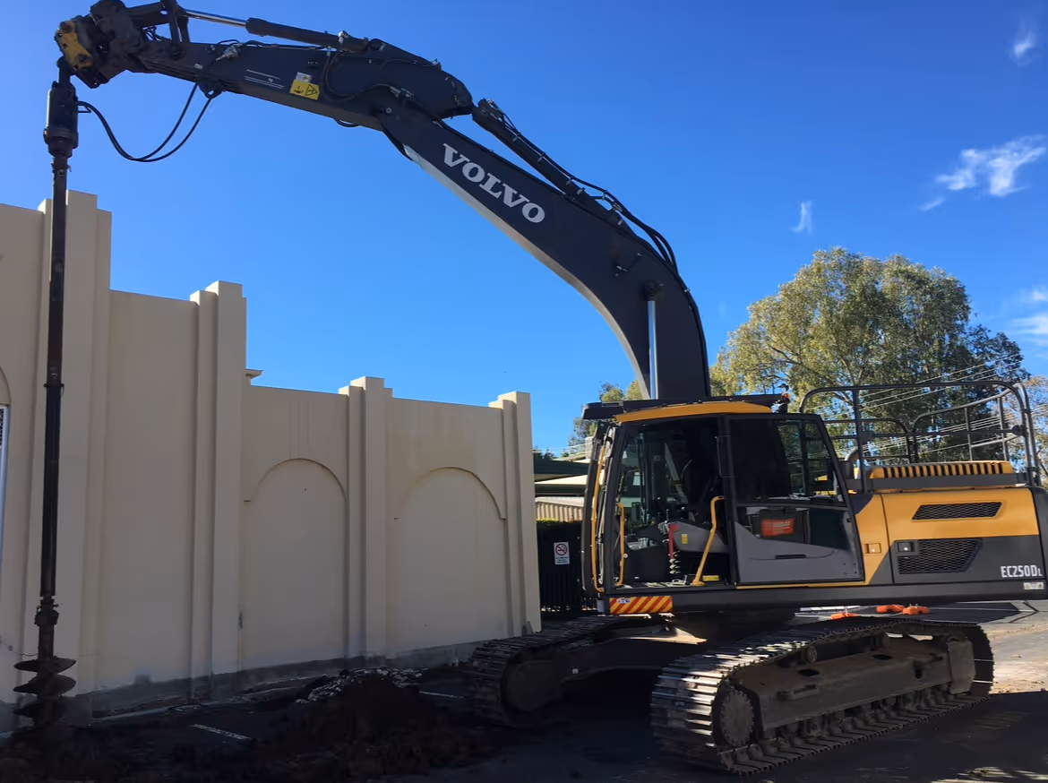 Yellow and black Volvo excavator with drilling attachment digging near a beige wall under clear blue sky.