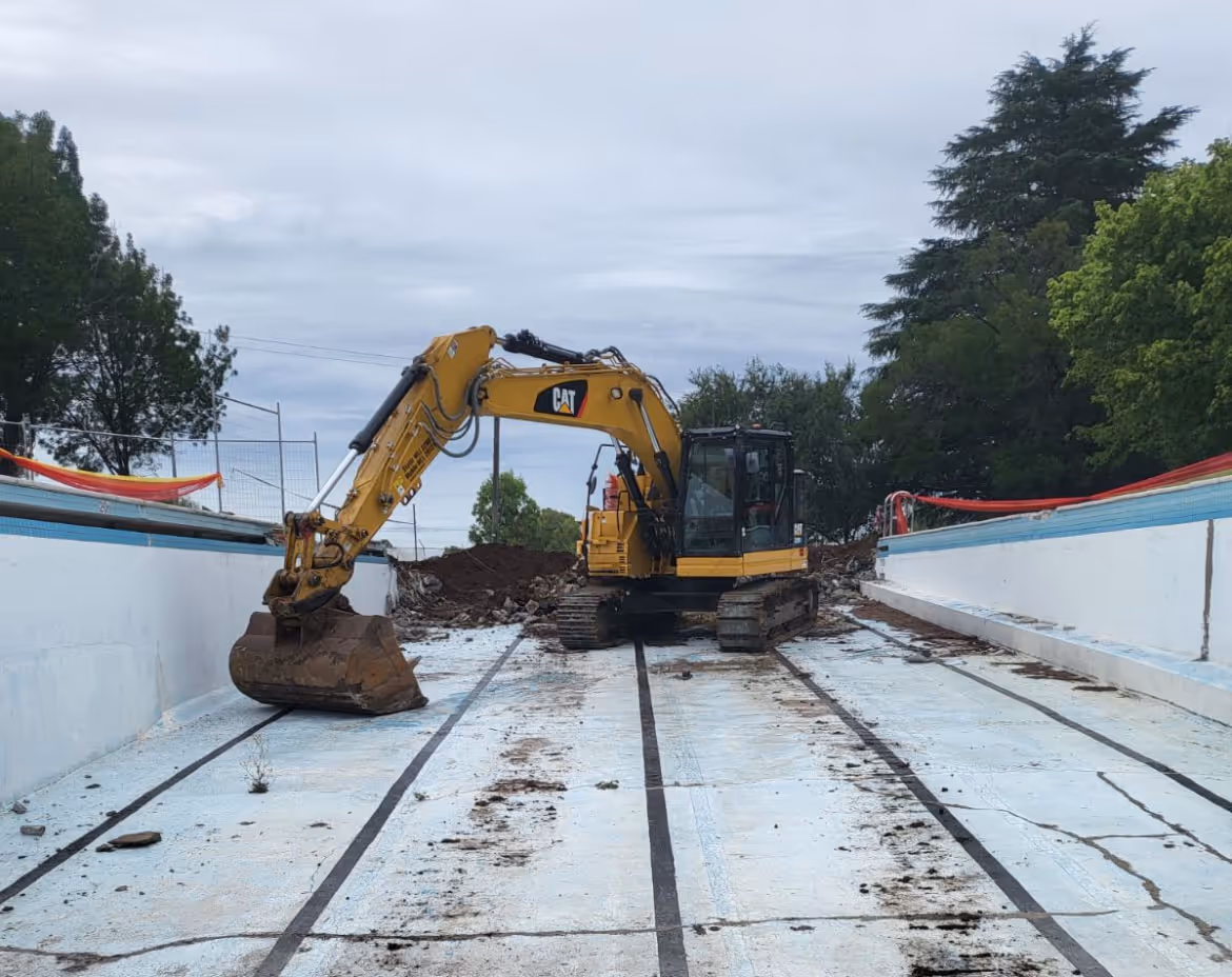 Yellow excavator inside an empty swimming pool with dirt and debris on the pool floor.