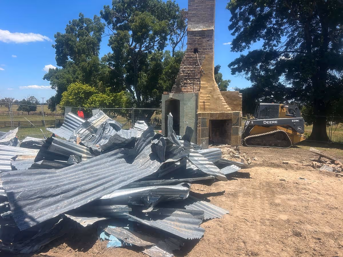Pile of corrugated metal sheets beside a partially demolished stone fireplace with a John Deere 329E compact track loader in the background.