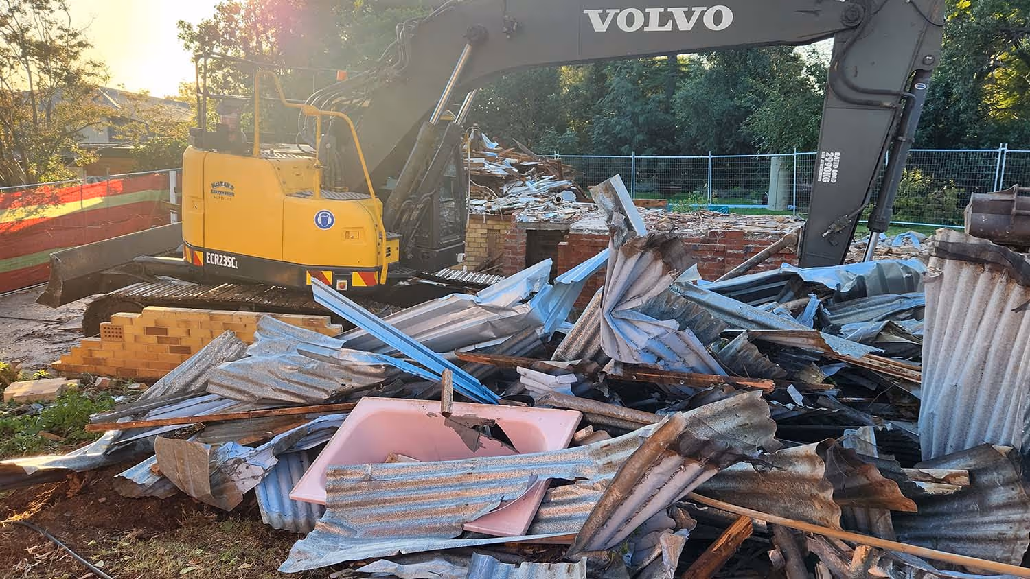 Yellow Volvo excavator at a demolition site with corrugated metal debris and a broken pink bathtub in front.