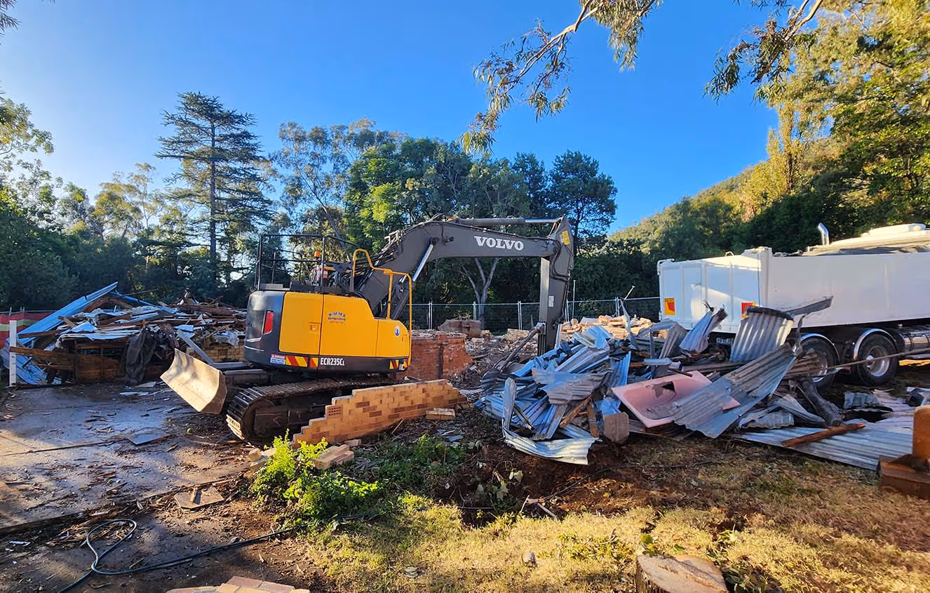 Yellow and black Volvo excavator with a bucket at a demolition site surrounded by debris and trees.