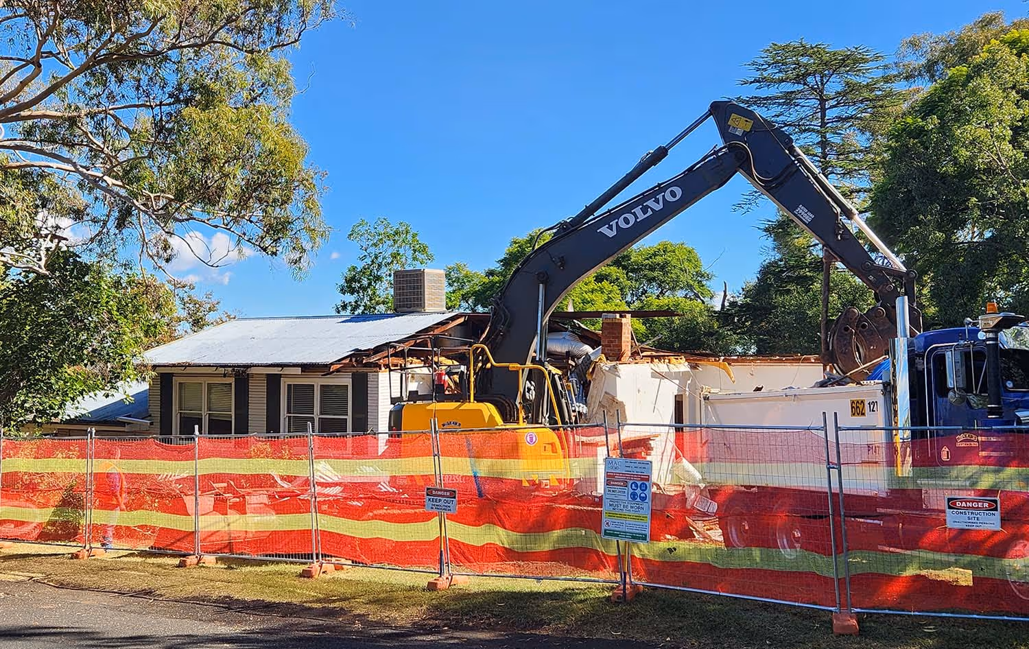 Demolition of a house with a Volvo excavator behind a red and yellow safety fence under a clear blue sky.