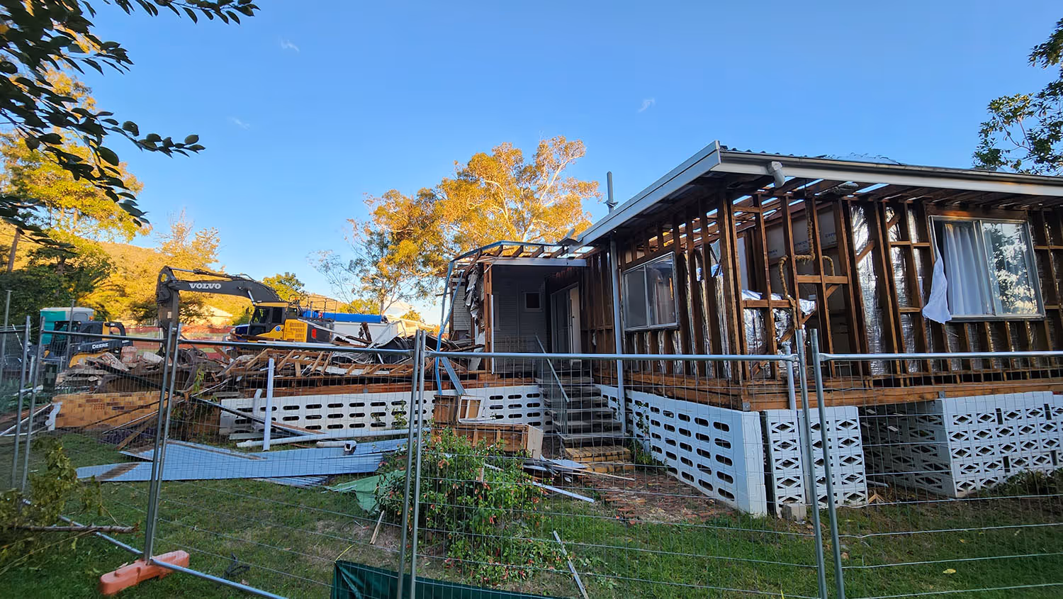 Partially demolished house with exposed wooden frame and debris inside construction fencing, with trees and a clear blue sky in the background.