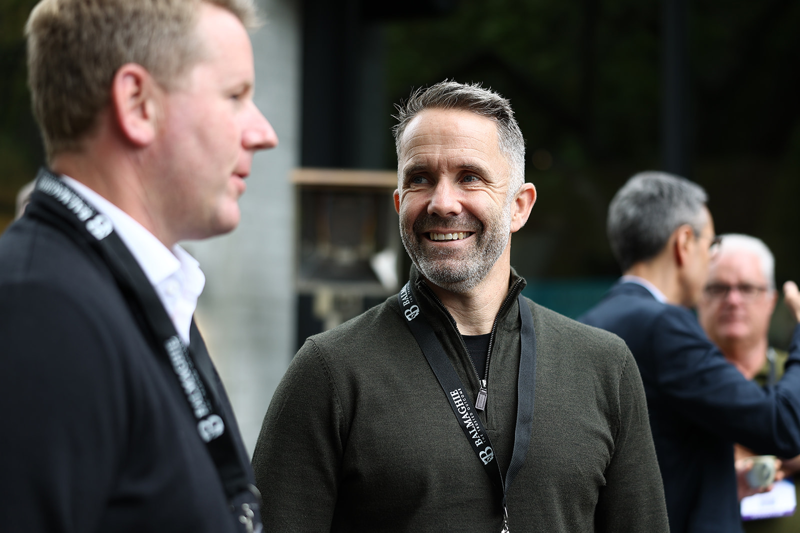 Two men wearing event lanyards talking outdoors, with other attendees behind them
