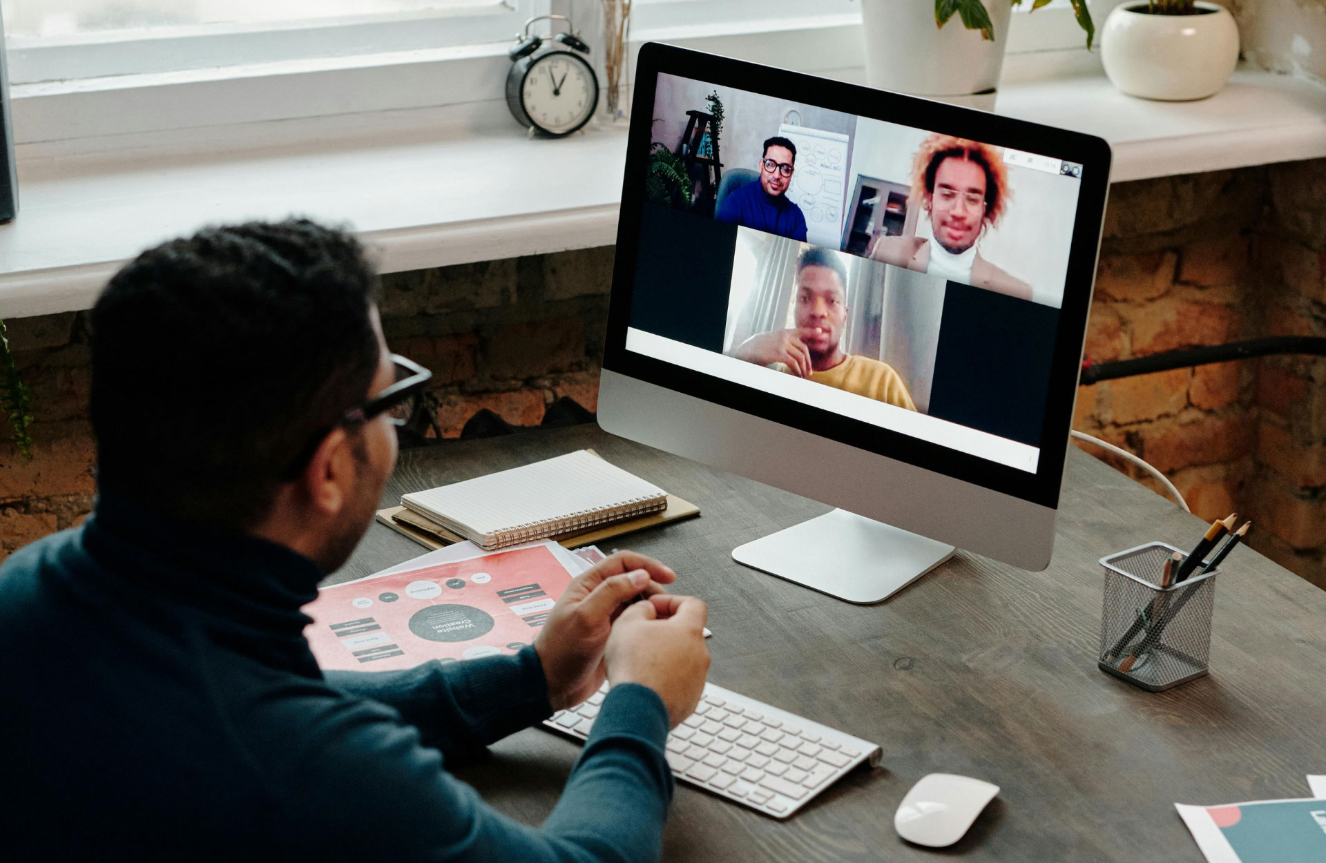 A man in a dark sweater participates in a video call on his desktop. Three people appear on the screen, suggesting a collaborative virtual meeting.