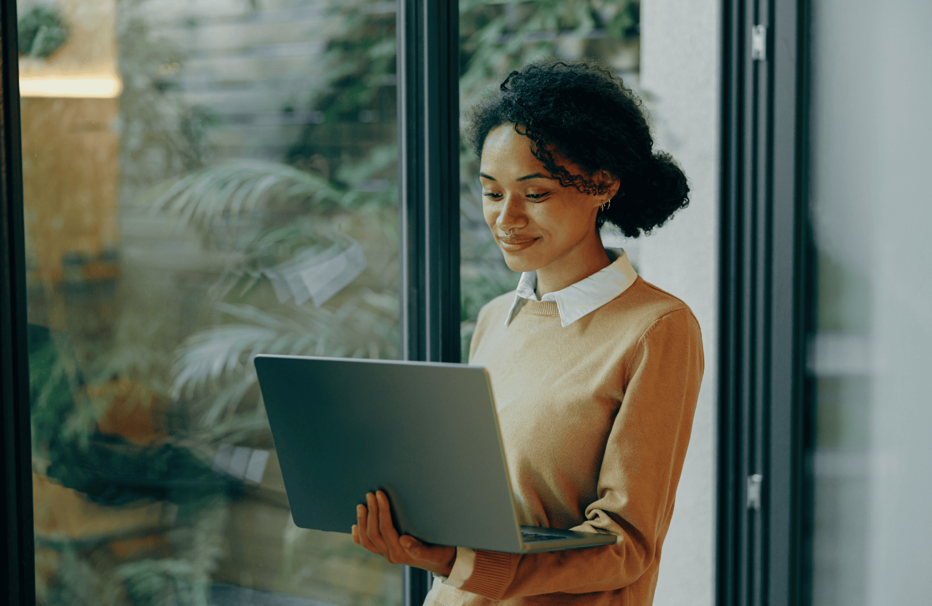 Smiling woman in a tan sweater holds a laptop near a window, with greenery outside. The atmosphere is calm and focused, suggesting productivity.