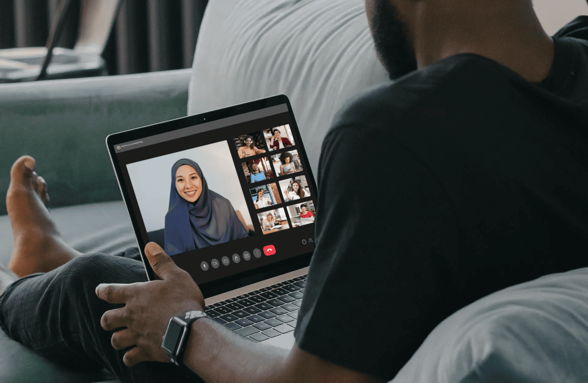 A person relaxes on a couch, participating in a video call on a laptop. The screen displays a smiling woman wearing a hijab with video thumbnails on the side.
