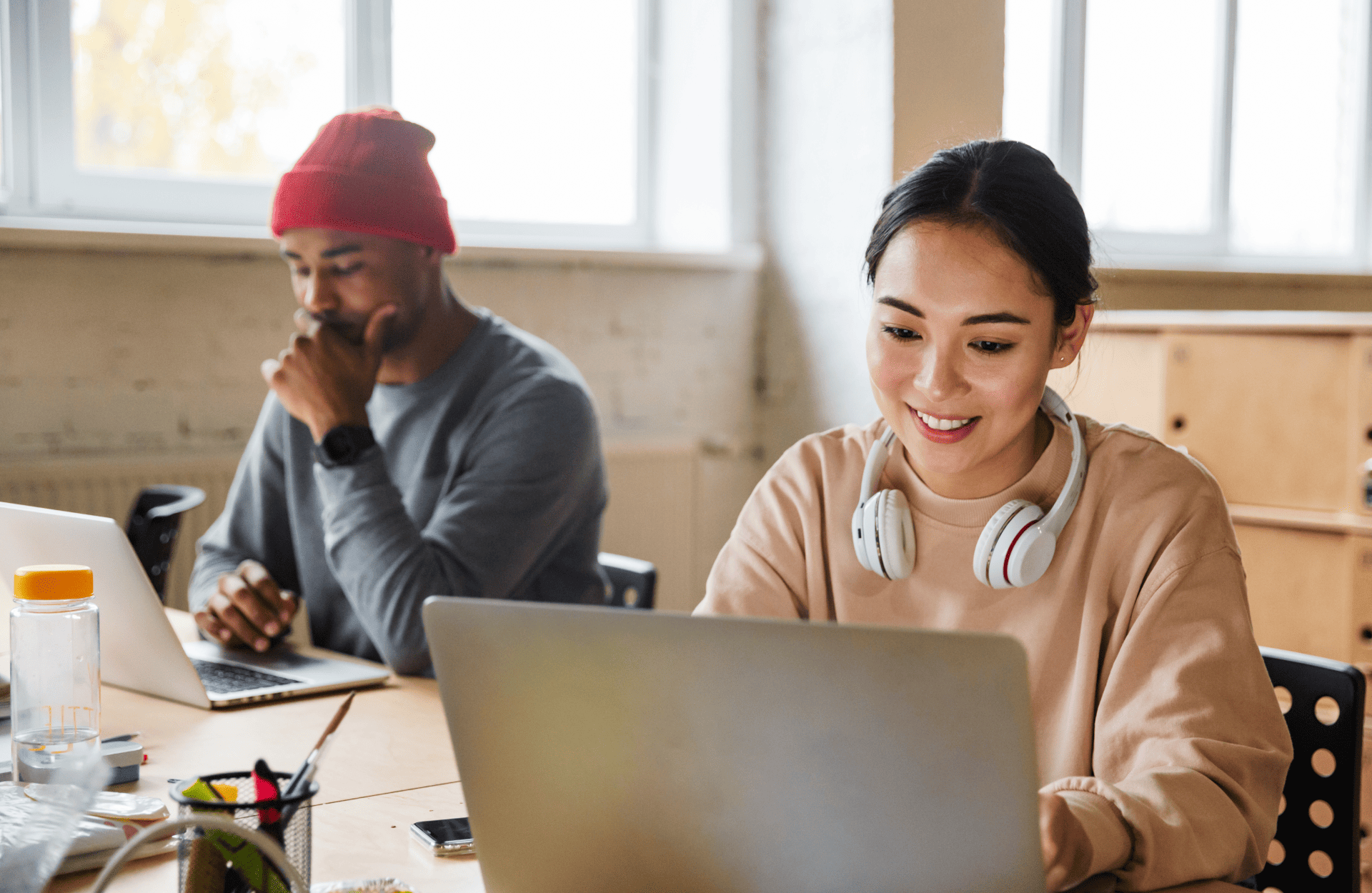 Two people working on laptops in a bright room. One wears a red beanie and looks thoughtful, while the other smiles with headphones around their neck.