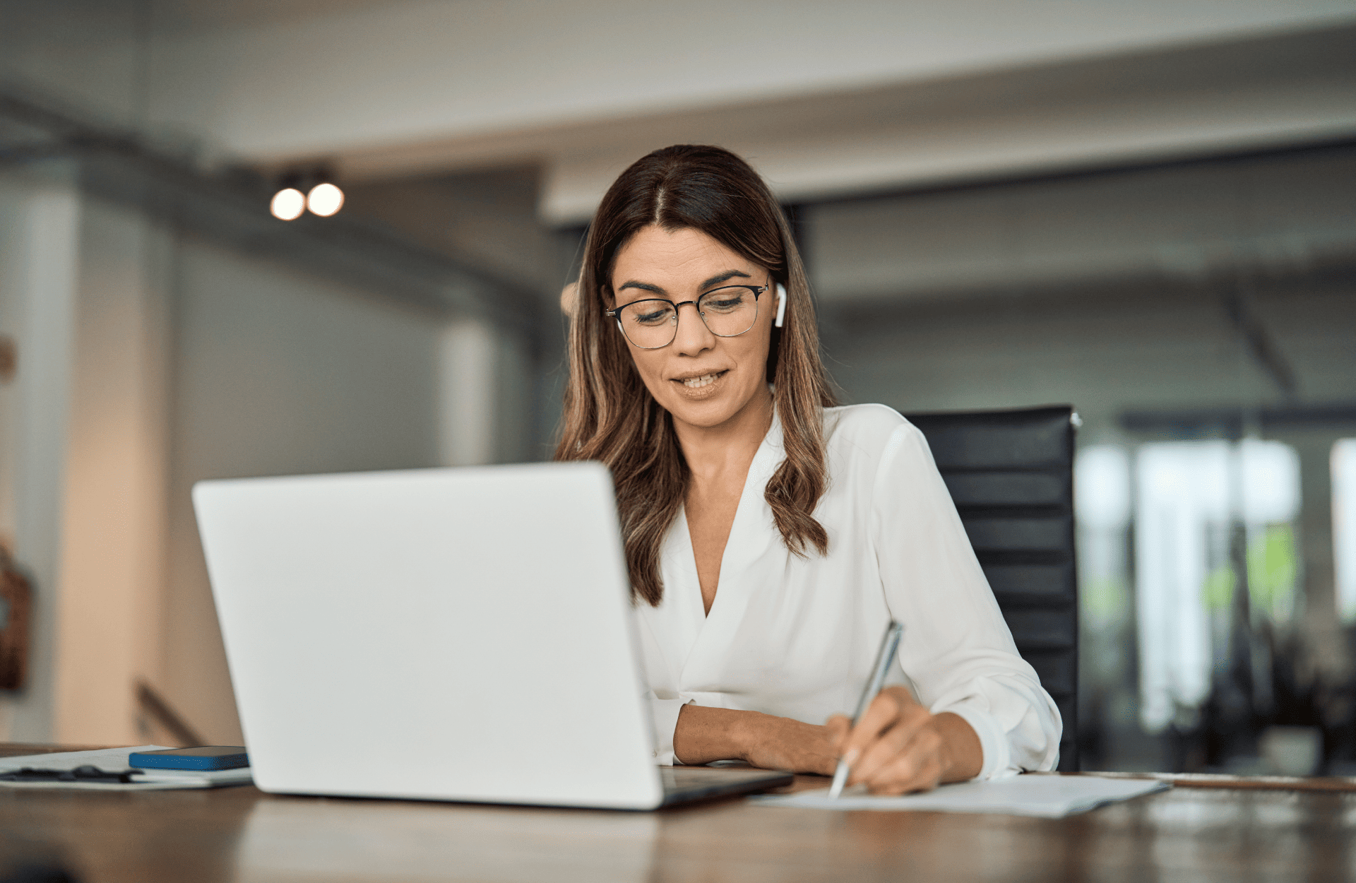 A woman in a white blouse sits at a desk with a laptop, writing on paper. She wears glasses and earbuds, conveying a focused and professional atmosphere.