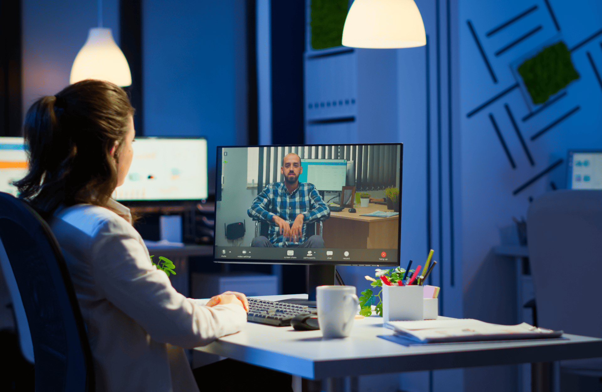 A woman in a home office is on a video call with a man on her computer screen. The room is dimly lit with a modern, professional atmosphere.