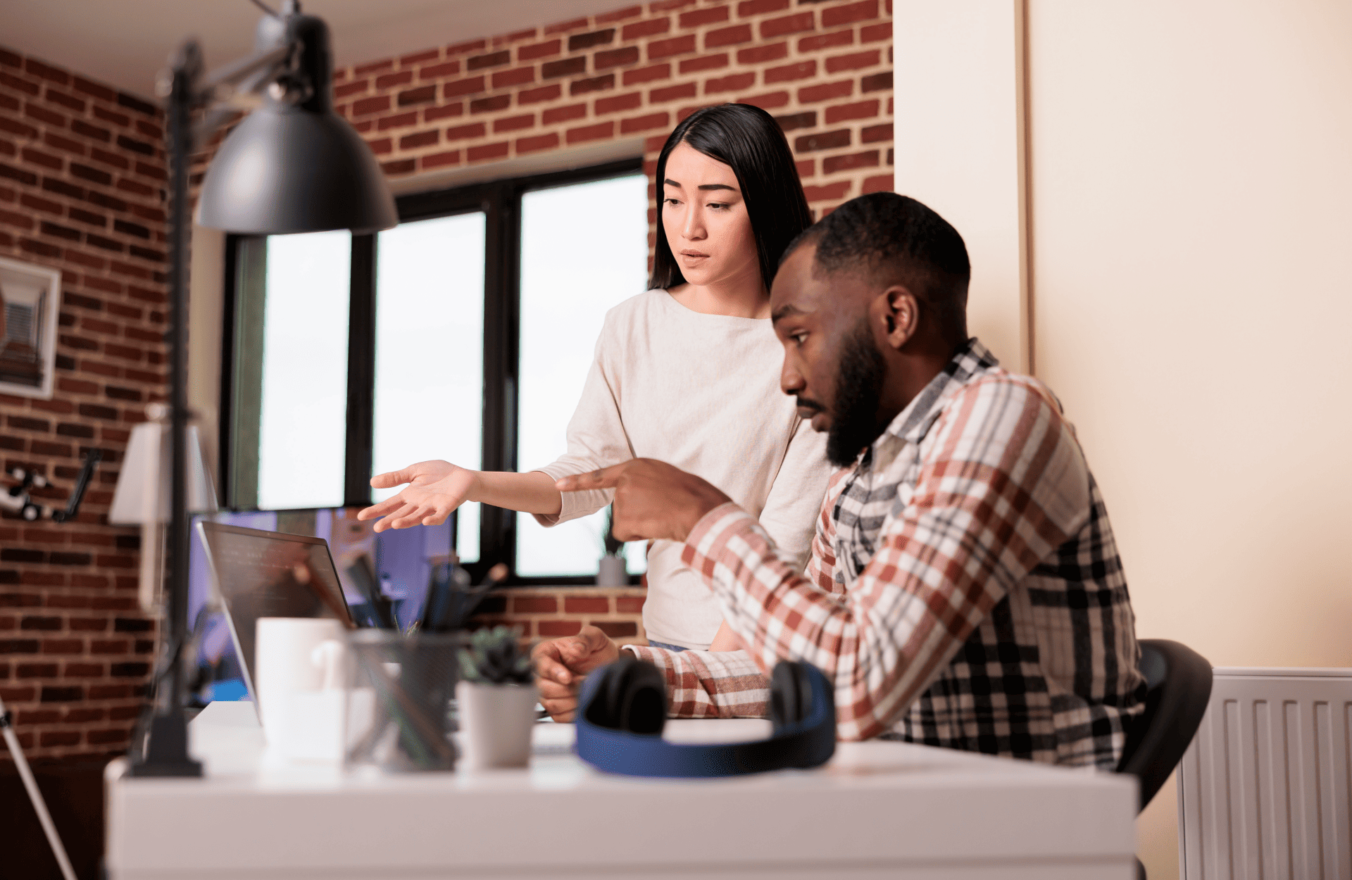 A woman stands beside a seated man at a desk, discussing something on a laptop. The room has a cozy brick wall, and the mood is focused and collaborative.