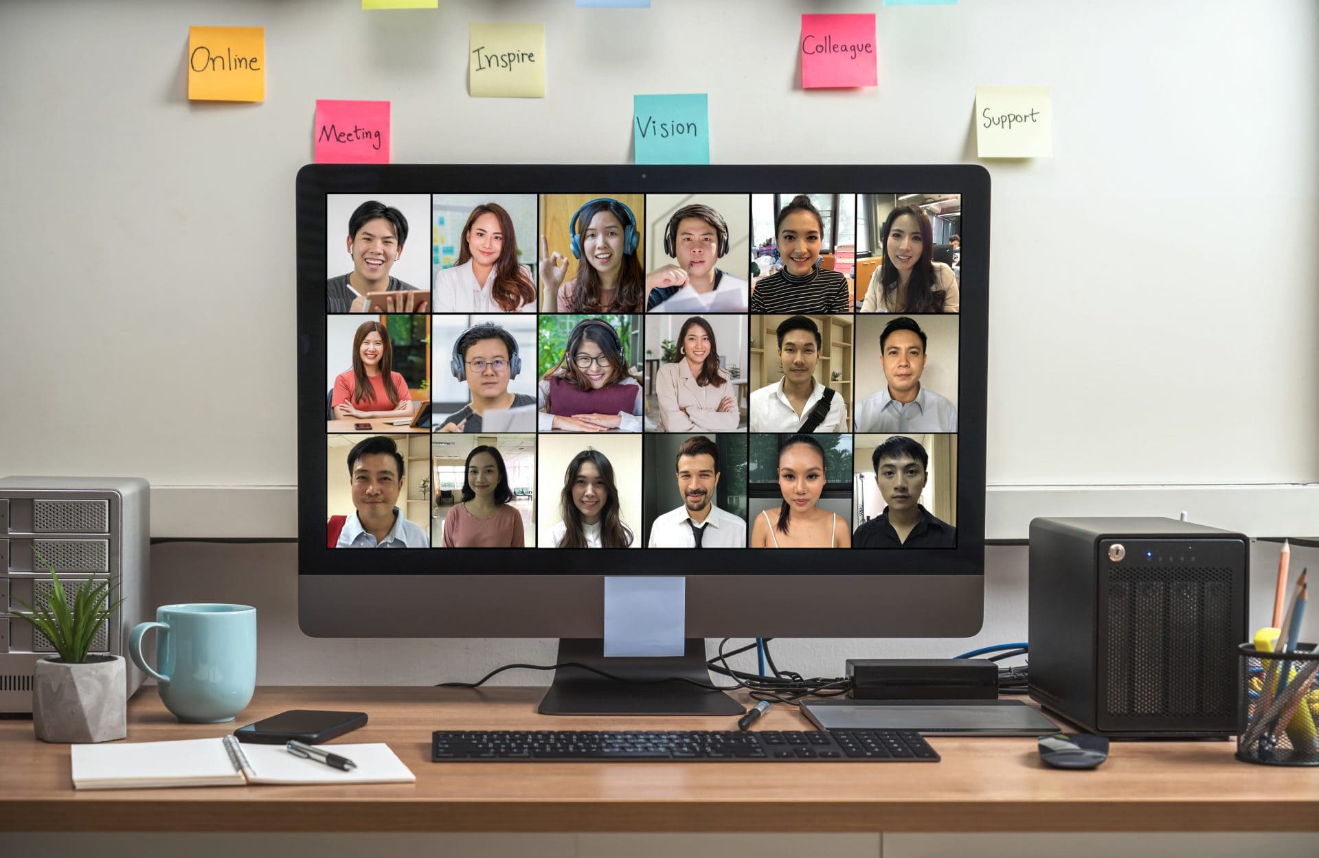 A computer screen shows a virtual meeting, with diverse participants smiling in a grid layout. Sticky notes with motivational words are on the wall.