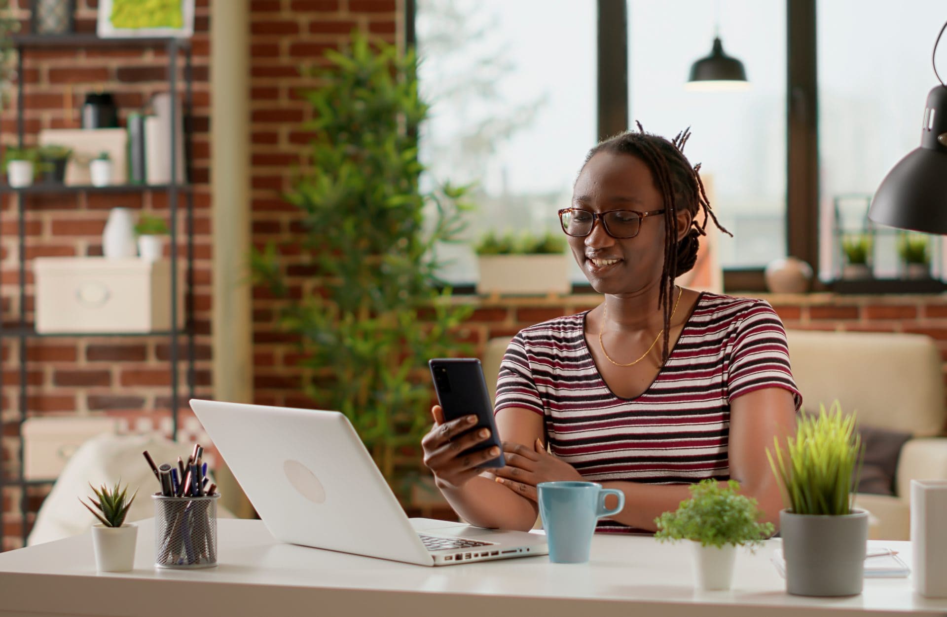 A person in glasses smiles while looking at a smartphone in a cozy home office. They sit at a white desk with a laptop, plants, and a blue mug.