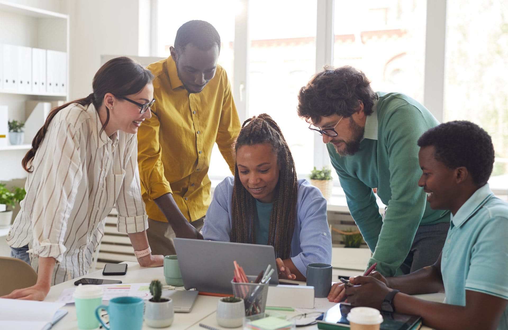 A diverse group of five people gather around a laptop in a bright office, collaborating with focus and enthusiasm. Papers and mugs are scattered on the table.