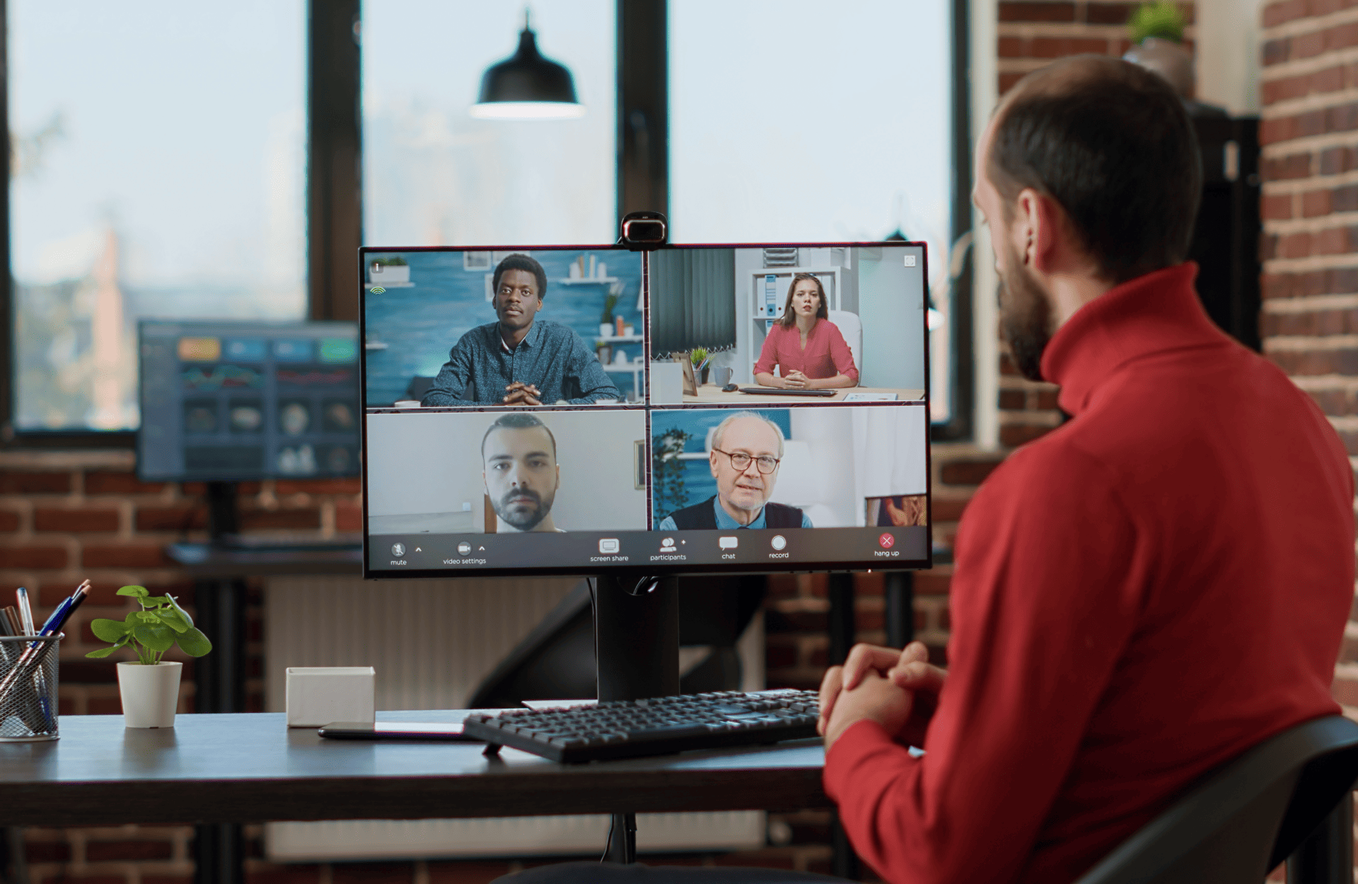 Man in a red sweater sits at a desk, engaged in a video call with four people on a monitor, in a modern brick-walled office setting.