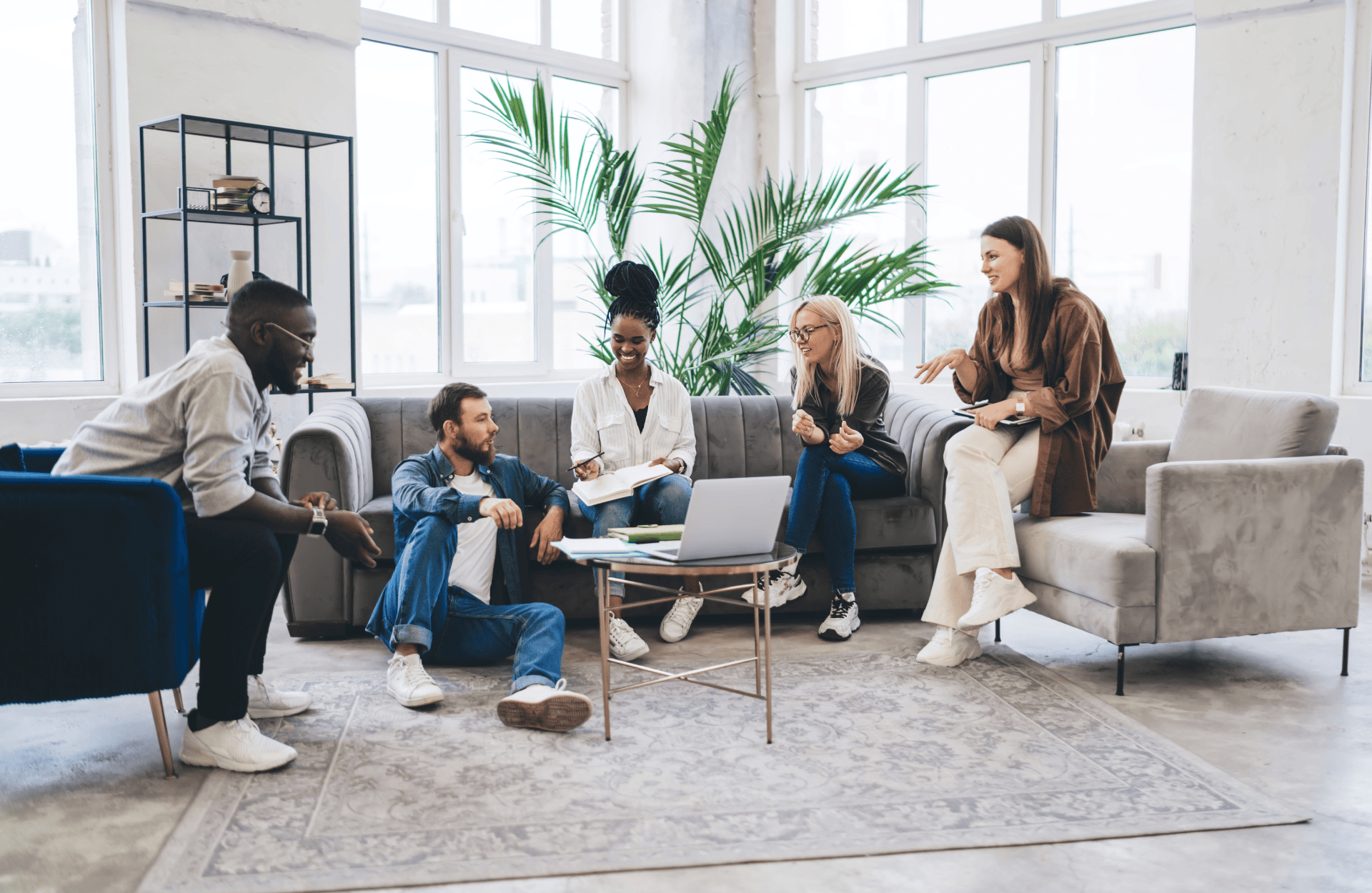 A diverse group of five people casually sit and converse in a modern, bright room with large windows. They're gathered around a laptop on a small table, suggesting a collaborative and relaxed atmosphere. A tall plant adds a touch of nature.
