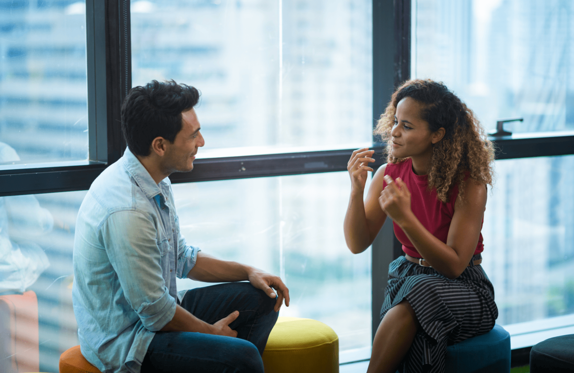 A man and woman engage in a lively conversation in a modern office. They're seated on colorful stools by a large window, with city buildings visible outside.