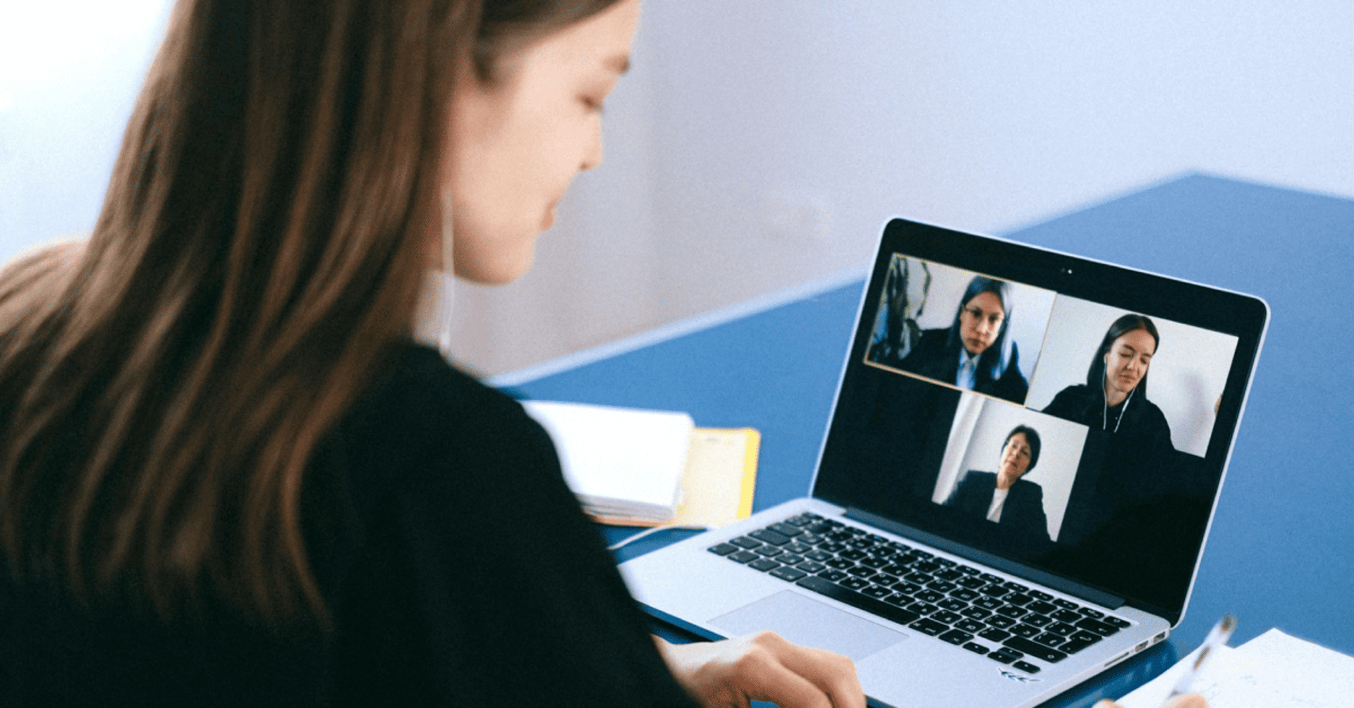 A woman participates in a video conference on a laptop, focusing on three people displayed on the screen. She's seated at a blue desk, writing notes.