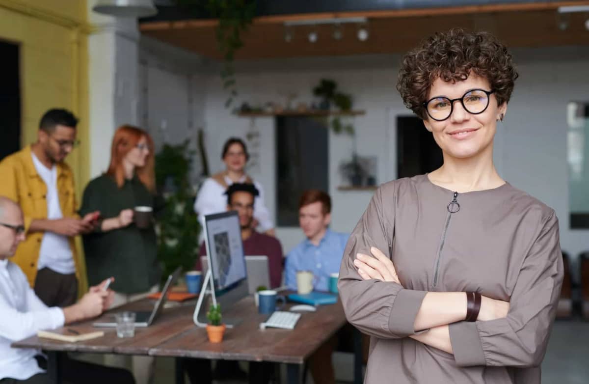 A confident person with curly hair and glasses stands smiling in the foreground of an office, with a diverse team working collaboratively in the background.