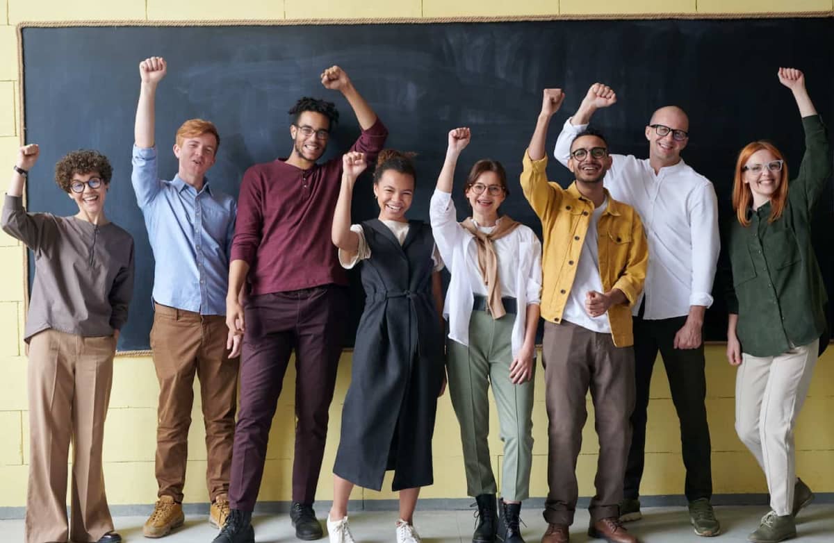 A diverse group of eight people stand with raised fists and cheerful expressions in front of a blackboard, conveying a sense of unity and celebration.