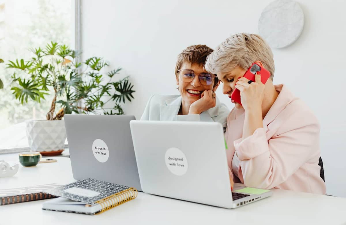 Two women smiling at a desk with laptops, one on a red phone. A potted plant and notebooks add a casual, friendly office vibe.