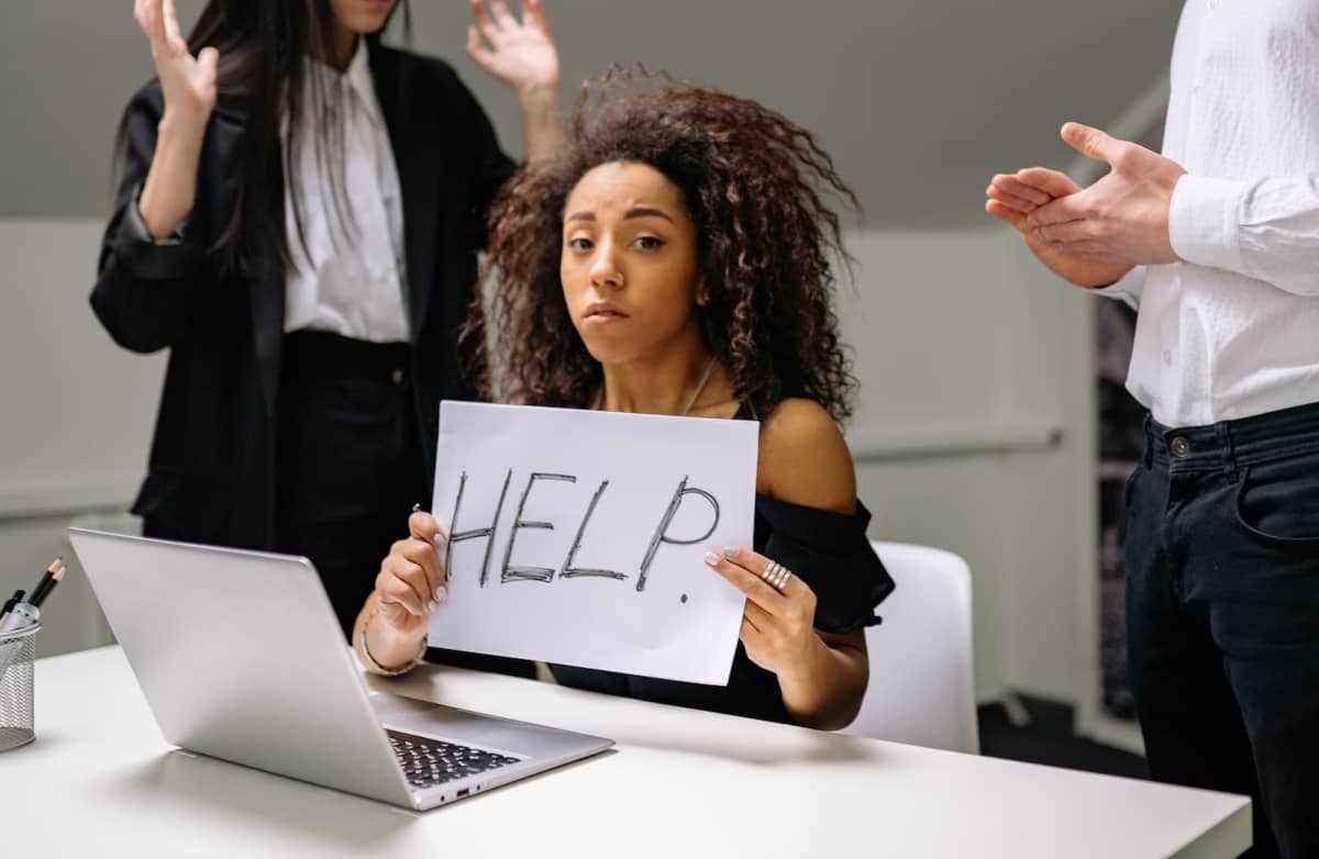 A woman sits at a desk holding a "HELP" sign, looking distressed. Two colleagues stand nearby gesturing, creating a tense office atmosphere.