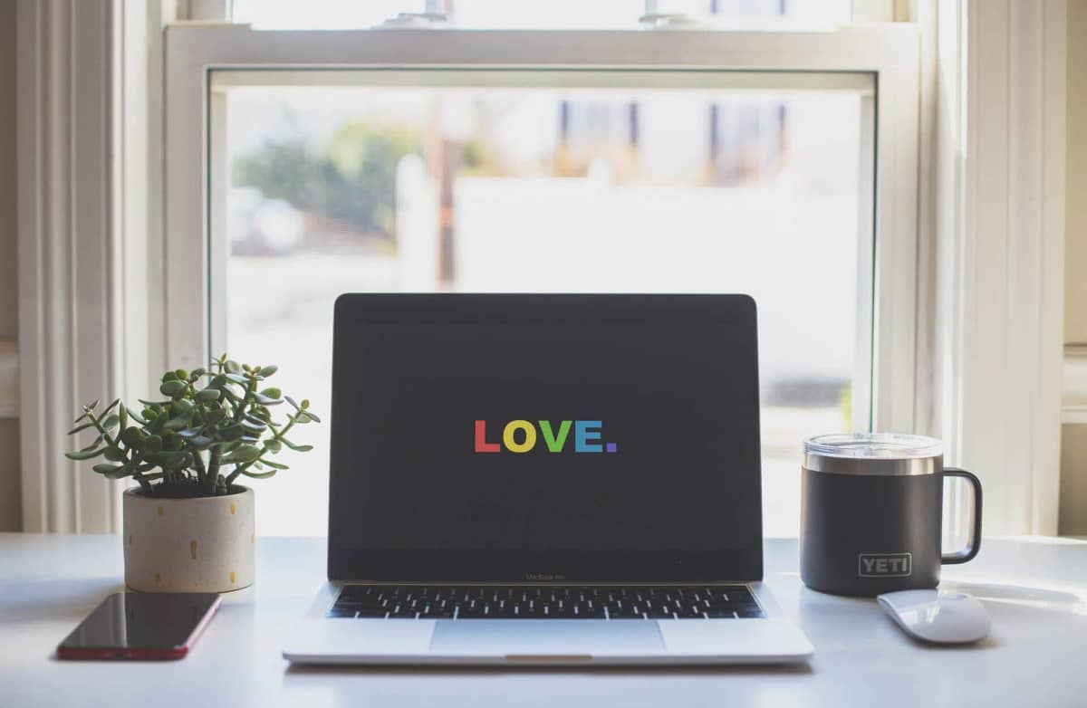 A laptop on a desk displays the word "LOVE" in rainbow colors. Beside it are a potted plant, a smartphone, a Yeti mug, and a mouse, set against a bright window.
