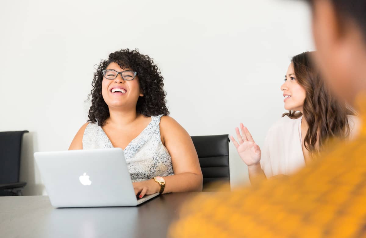 Two people in a meeting room, one laughing joyfully at a desk with a laptop, the other gesturing while talking, conveying a positive atmosphere.