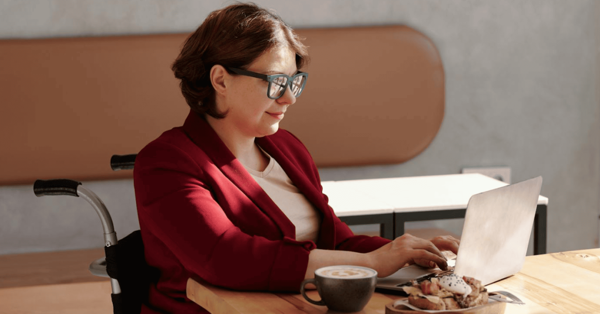 A woman in a red blazer, seated in a wheelchair, works on a laptop at a table. She appears focused, with a coffee and pastries nearby, in a cozy setting.