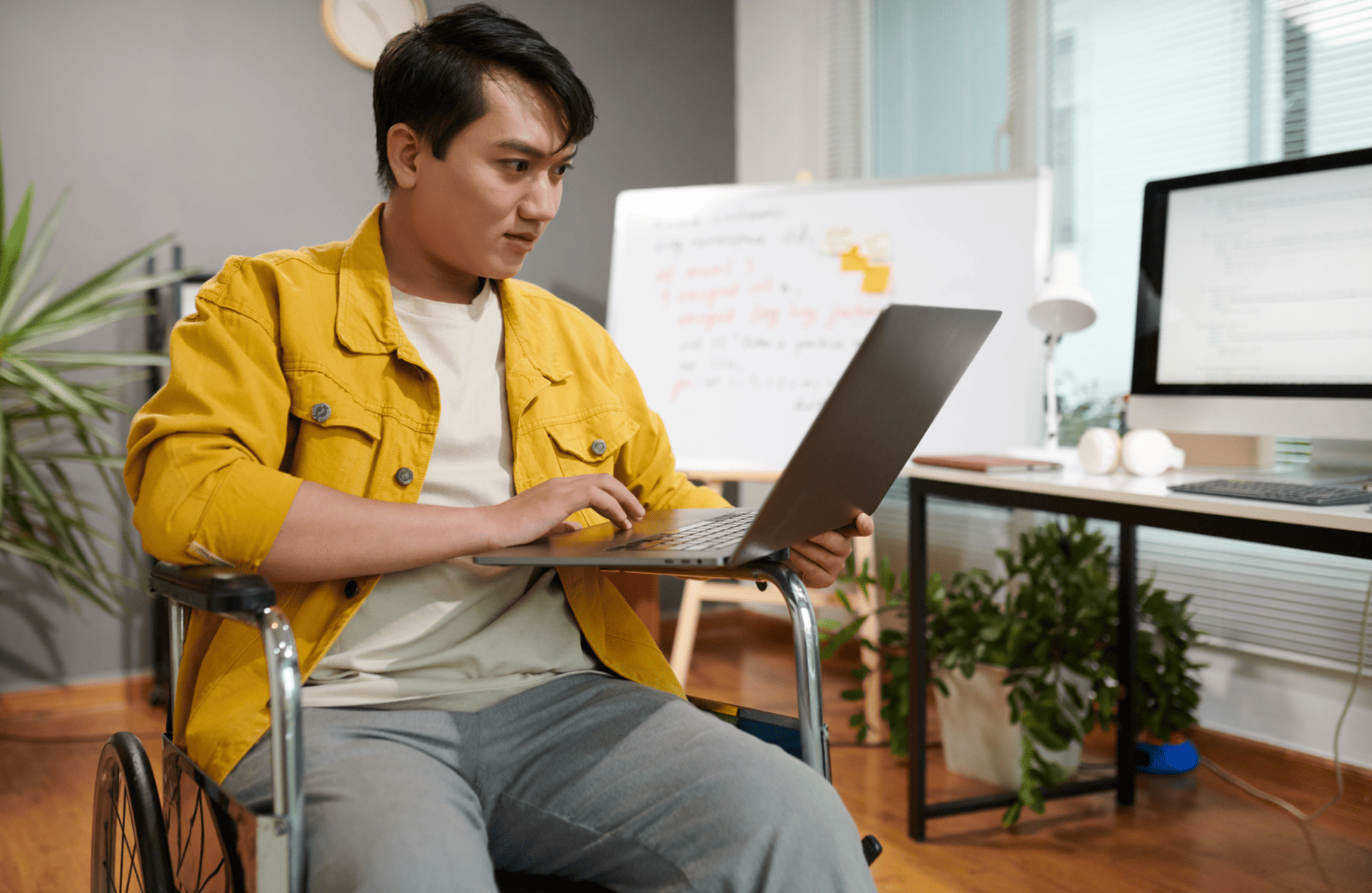 A person in a yellow jacket works on a laptop in a wheelchair. The room has a modern, cozy vibe with a chalkboard, computer, plants, and wooden flooring.