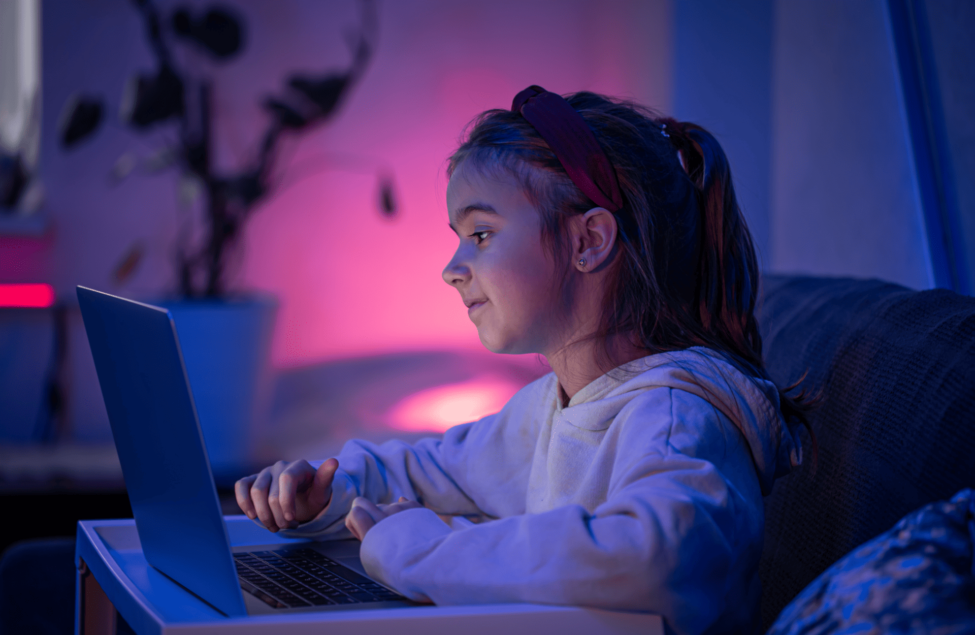 A young girl in a white hoodie and headband sits on a couch, focused on a laptop. The room is warmly lit with pink and blue tones, creating a cozy ambiance.
