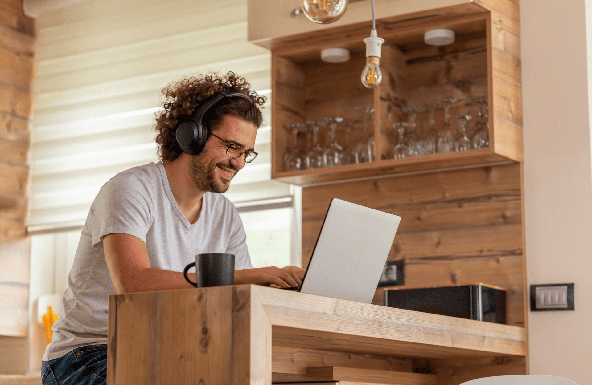 Man with curly hair sits at a wooden kitchen counter, wearing headphones and smiling at a laptop. A black mug is beside him, and shelves with glasses are in the background.