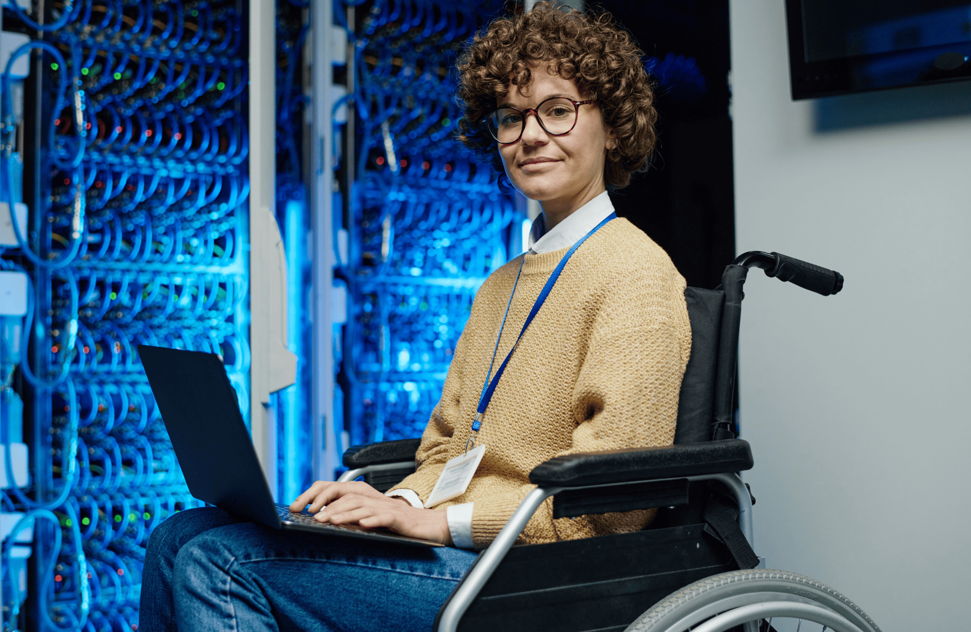 A person in a wheelchair works on a laptop in a server room, surrounded by glowing blue lights. They wear glasses and a lanyard, exuding confidence.