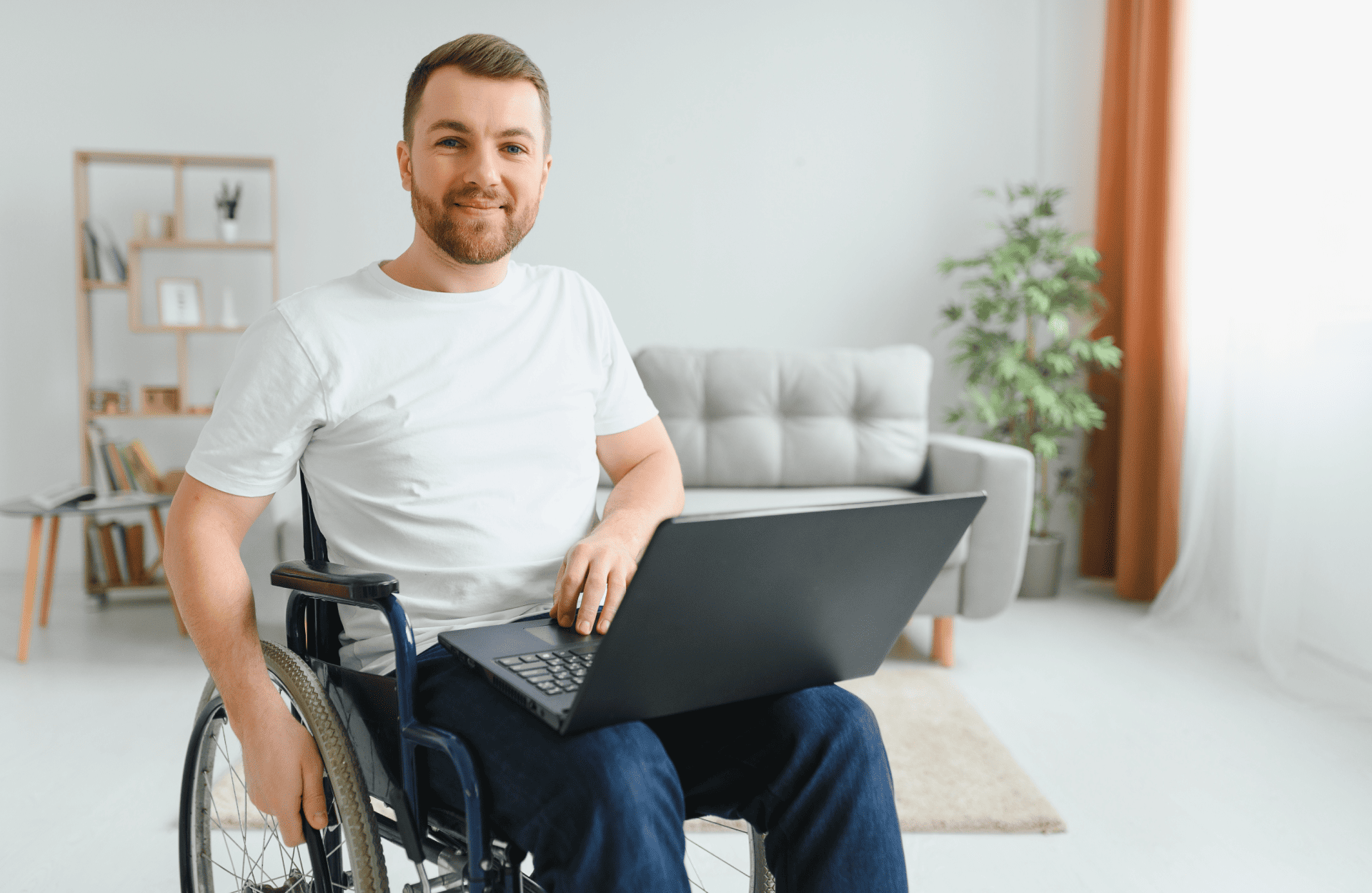 A smiling man in a wheelchair uses a laptop at home. The bright room has a grey sofa, a plant, and bookshelves, creating a cozy, productive atmosphere.