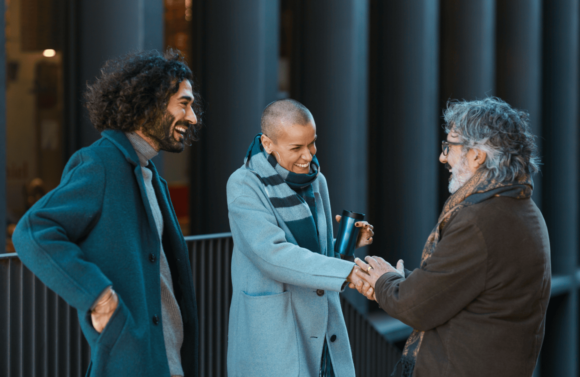 Three people in coats, two men and a woman, share a joyful conversation outdoors. They are smiling, with one shaking hands. The mood is warm and friendly.