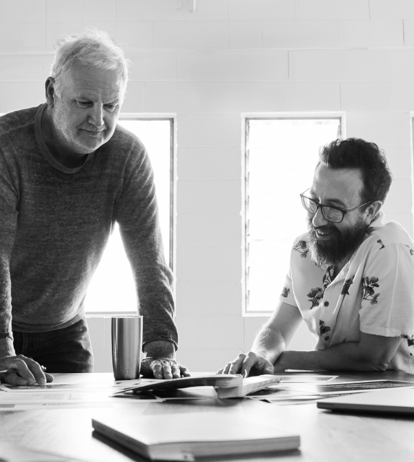 Two men collaborating over documents at a table, one seated smiling, the other standing and leaning in.