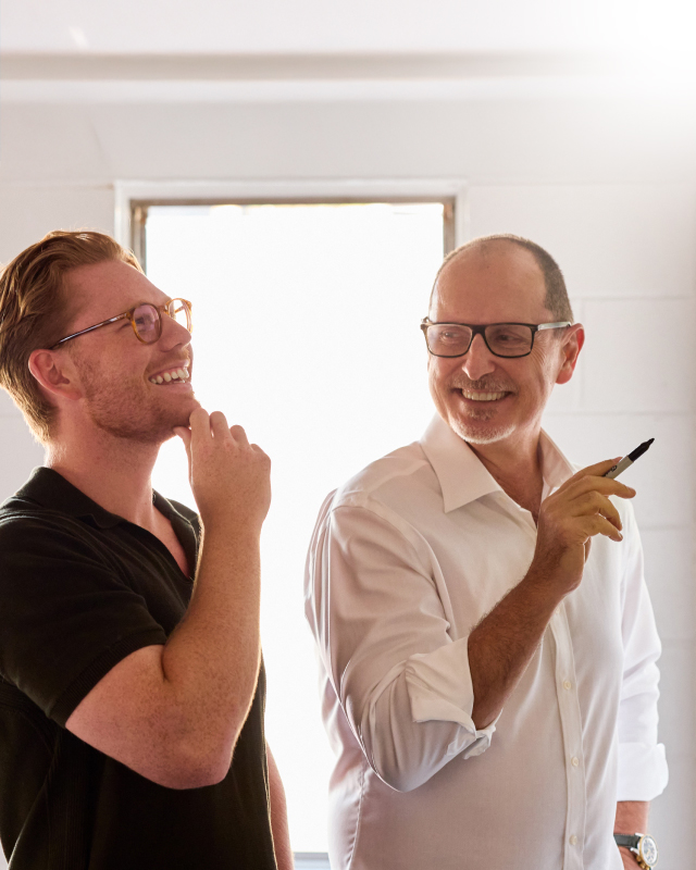 Two men smiling and looking at each other, one holding a marker, in a bright room.
