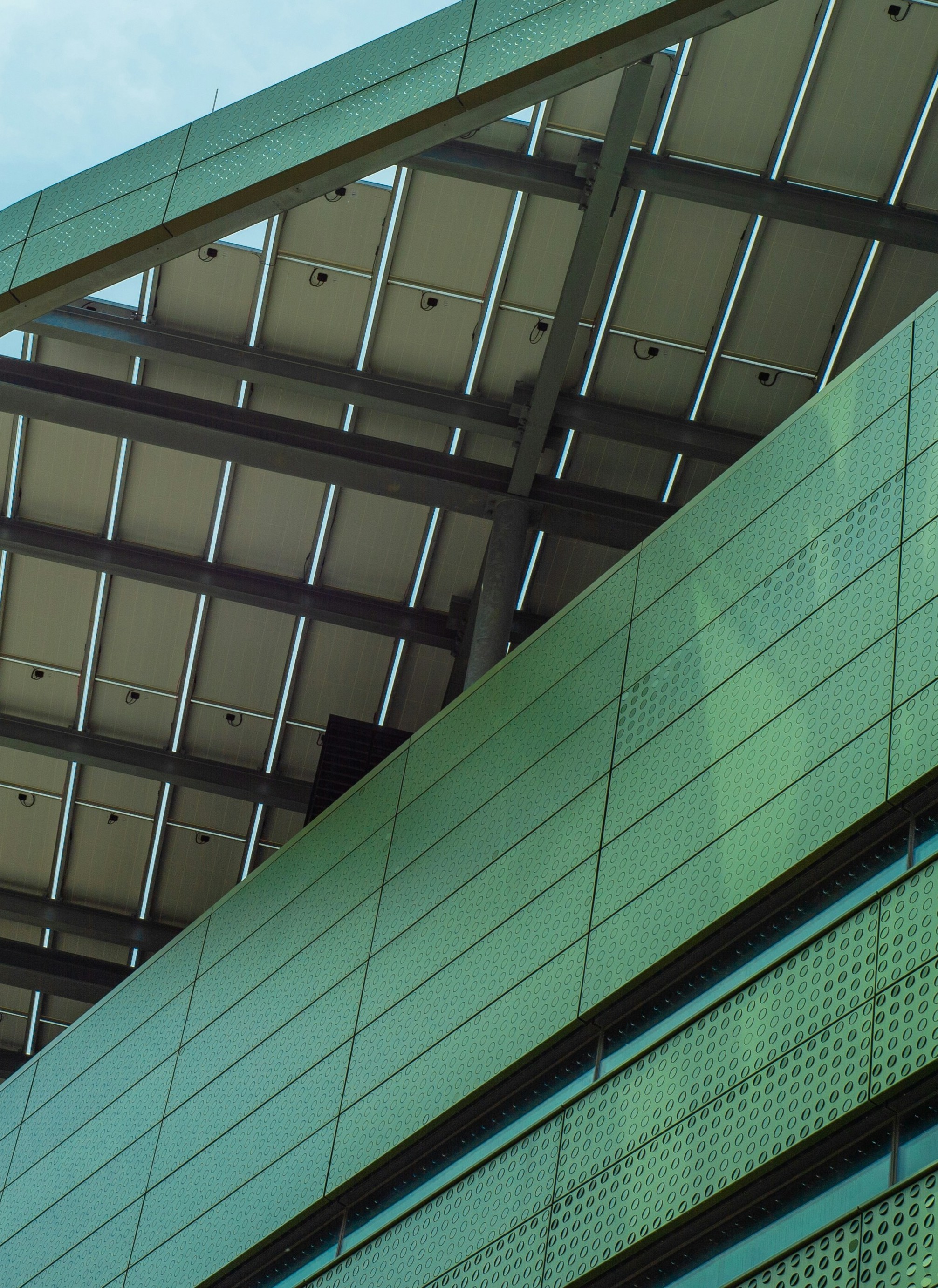 Green textured building facade with a metal framed roof structure above against a cloudy sky.