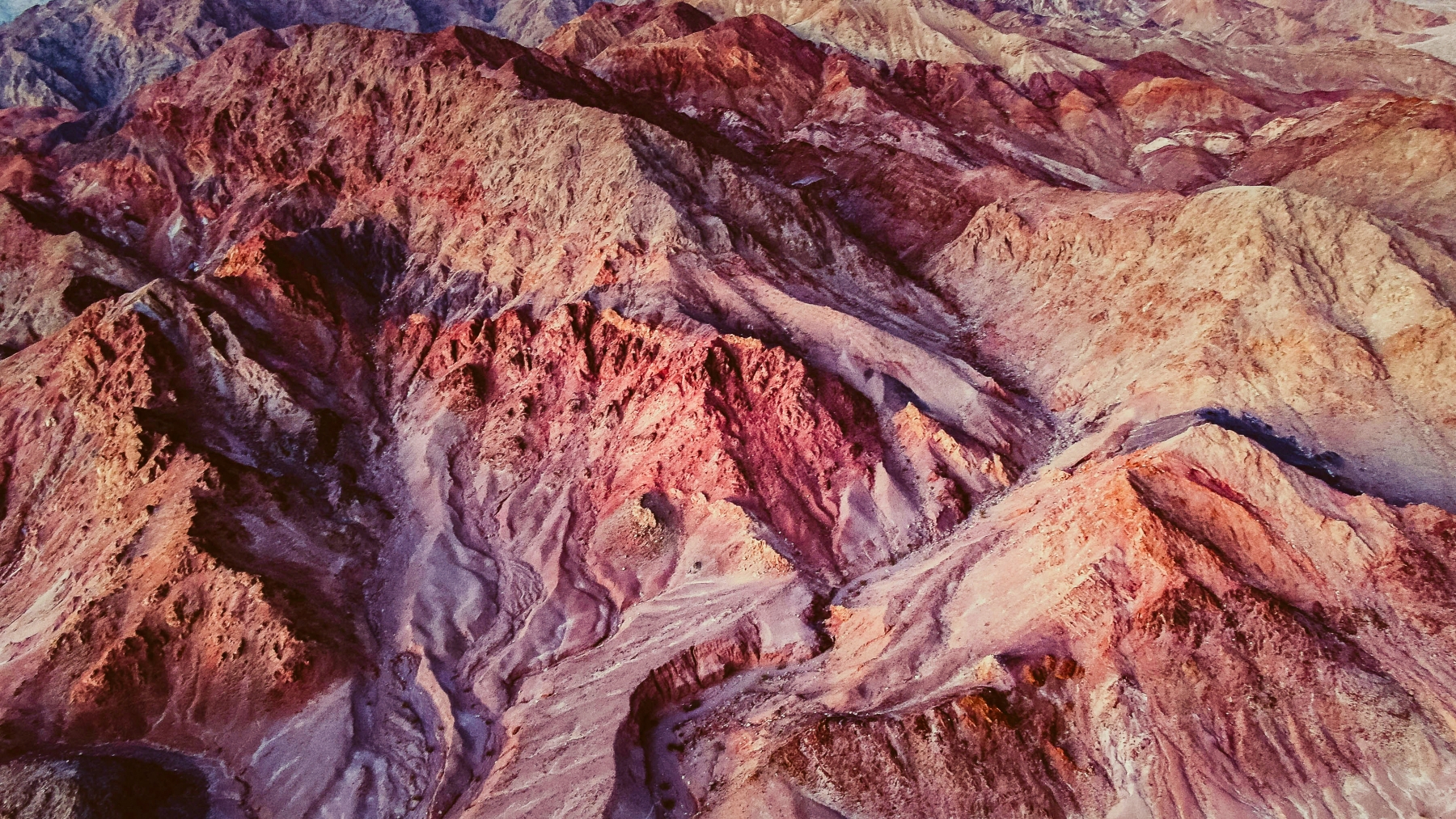 Aerial view of rugged desert mountains with reddish and brown hues and eroded ridges.
