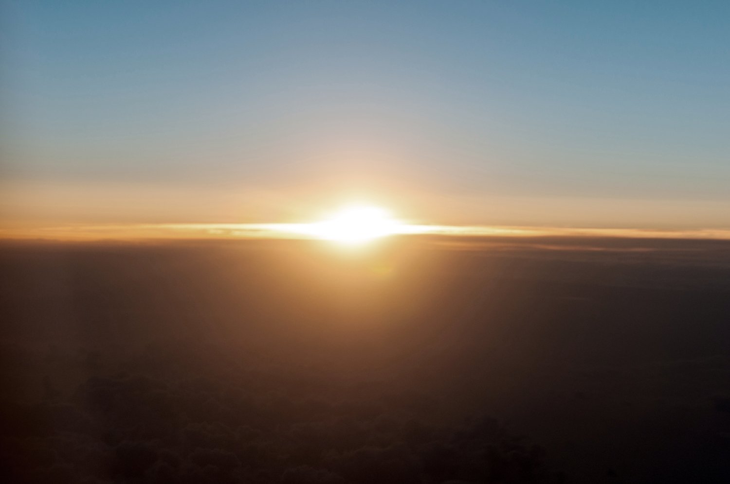 Bright sun setting over a horizon with a clear blue sky above and dark landscape below.