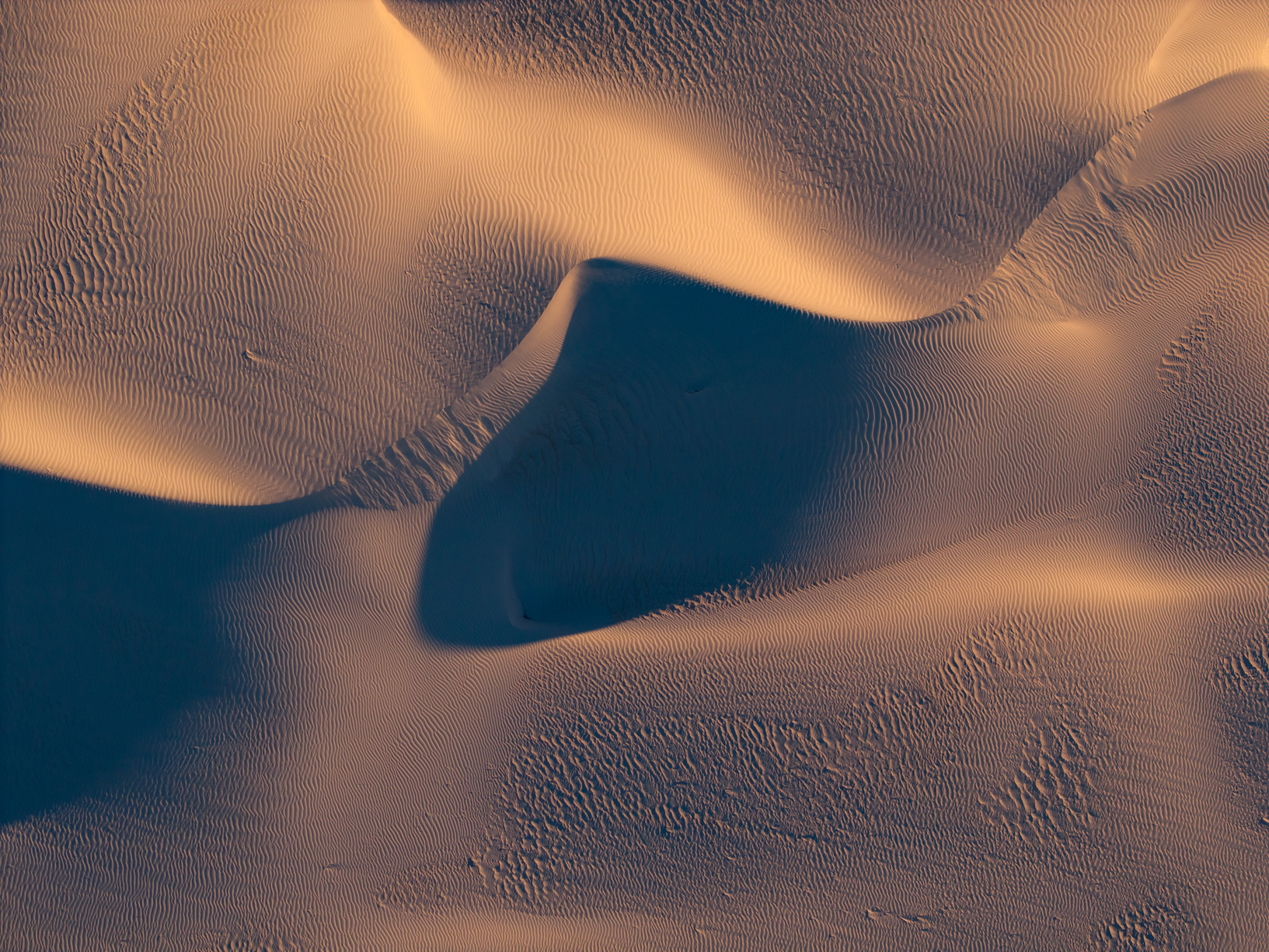 Aerial view of sand dunes with shadows highlighting their textured, rippled patterns in golden light.