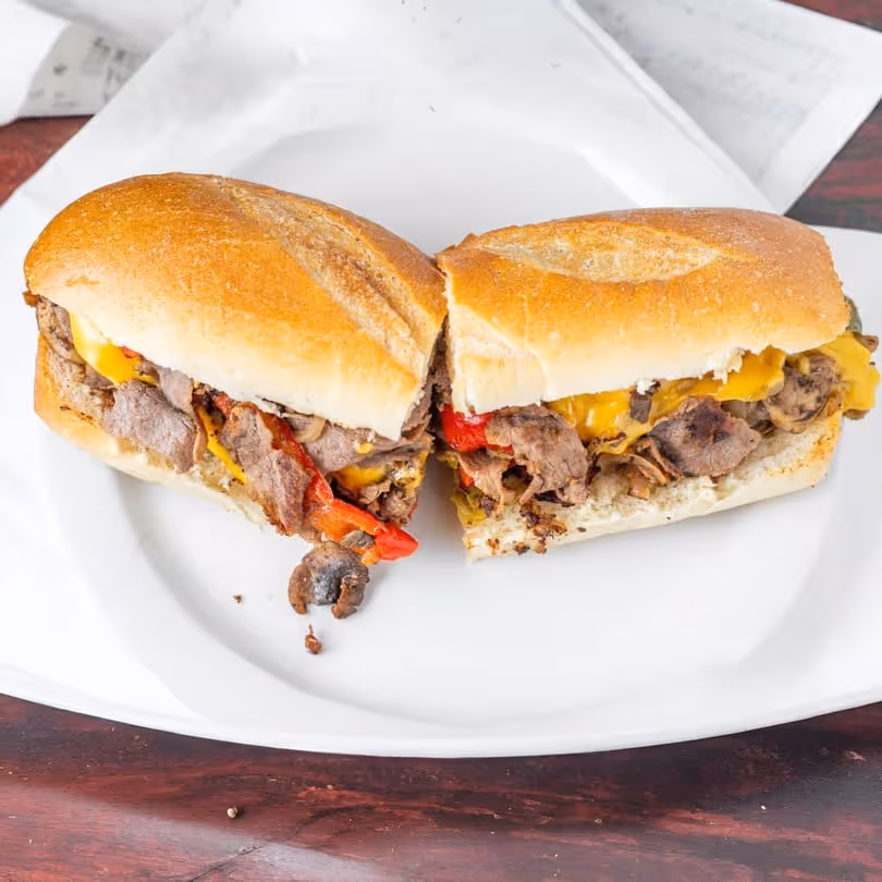 Philly cheesesteak sandwich cut in half with melted cheese, sliced steak, mushrooms, and red peppers on a white plate.